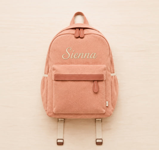 A dusty rose canvas backpack with a front zippered pocket and the name 'Sienna' embroidered in white script on the front, displayed flat on a light wood background.