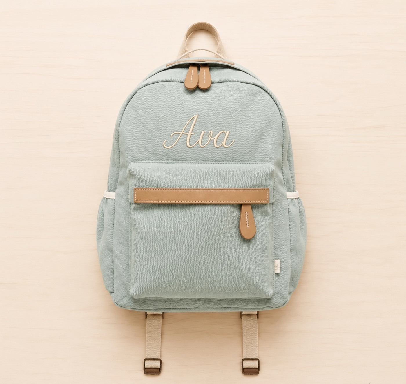 A sage green canvas backpack with tan leather accents and the name 'Ava' embroidered in gold on the front, displayed flat on a light wood background.