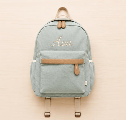 A sage green canvas backpack with tan leather accents and the name 'Ava' embroidered in gold on the front, displayed flat on a light wood background.