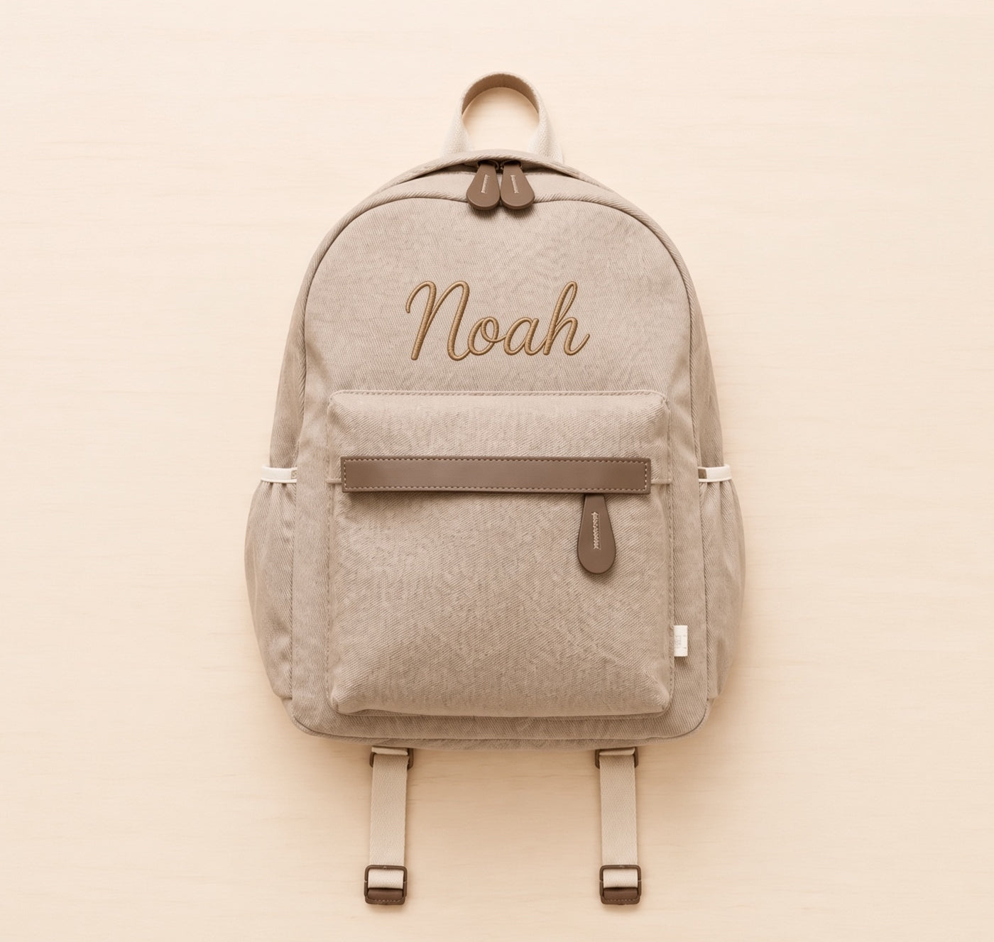 A light beige backpack with brown accents, featuring the name 'Noah' embroidered in brown cursive on the front pocket. The backpack is displayed flat against a light beige background.