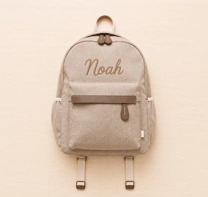 A light beige backpack with brown accents, featuring the name 'Noah' embroidered in brown cursive on the front pocket. The backpack is displayed flat against a light beige background.