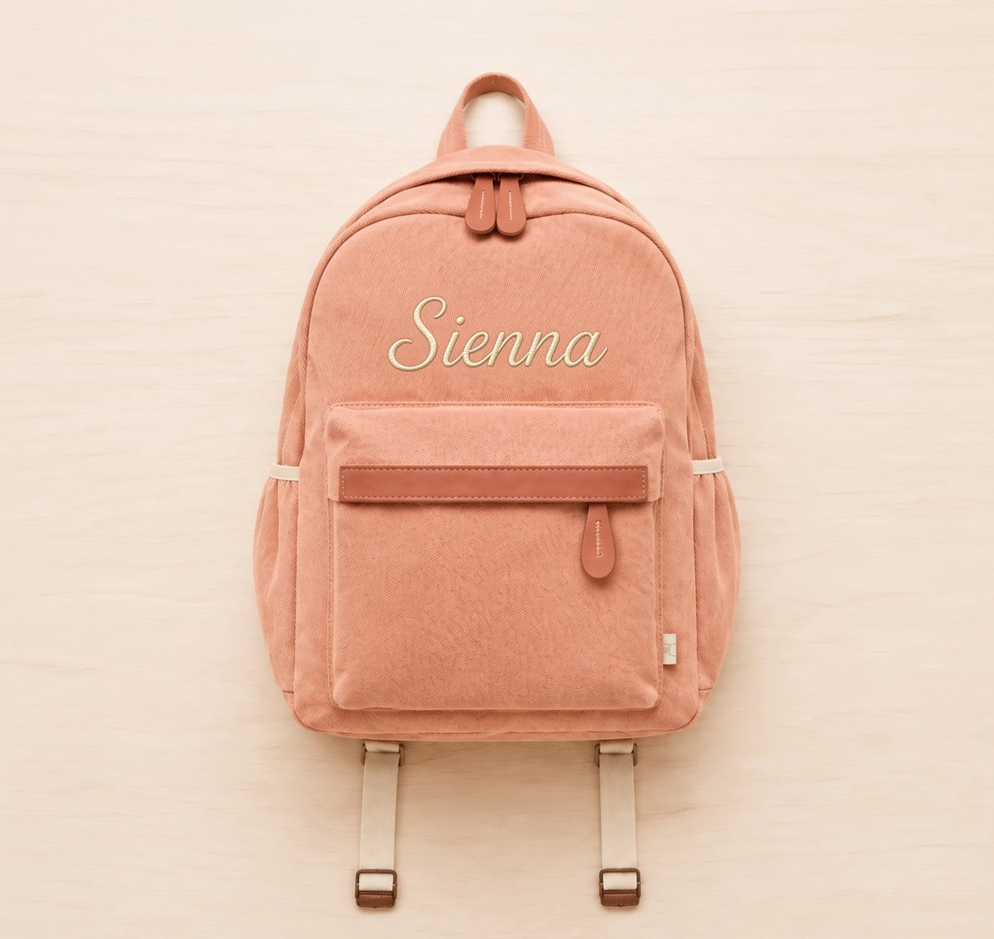 A dusty rose canvas backpack with a front zippered pocket and the name 'Sienna' embroidered in white script on the front, displayed flat on a light wood background.