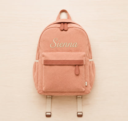 A dusty rose canvas backpack with a front zippered pocket and the name 'Sienna' embroidered in white script on the front, displayed flat on a light wood background.