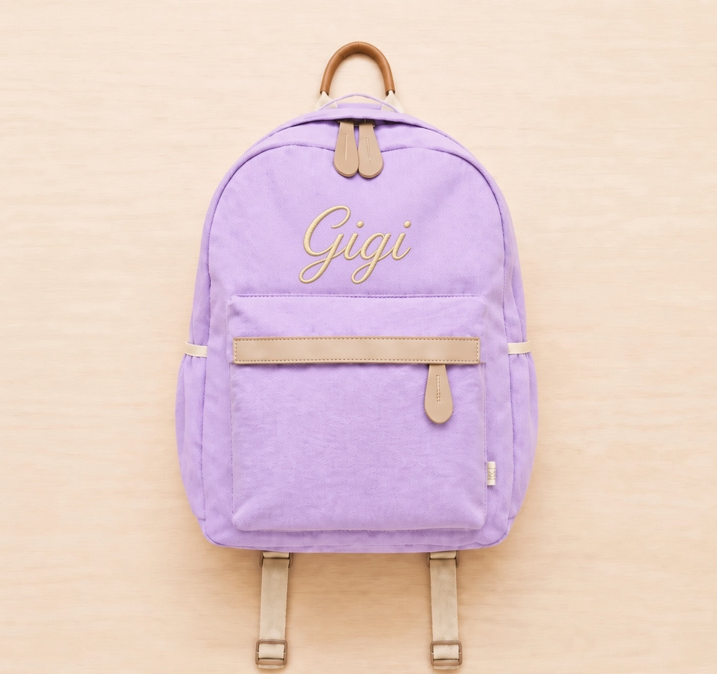 A lavender purple canvas backpack with tan leather-like trim and straps, featuring the name 'Gigi' embroidered in gold cursive on the front pocket, set against a light wood background.
