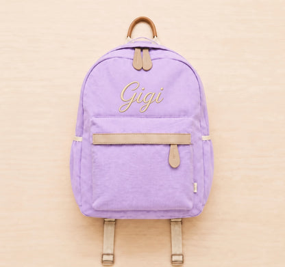 A lavender purple canvas backpack with tan leather-like trim and straps, featuring the name 'Gigi' embroidered in gold cursive on the front pocket, set against a light wood background.